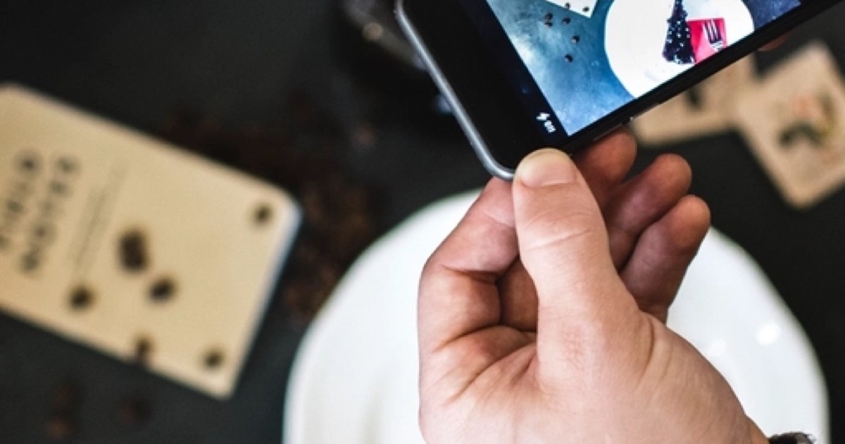 A hand holding a smartphone over a white plate on a dark table, framing the plate on the phone screen.