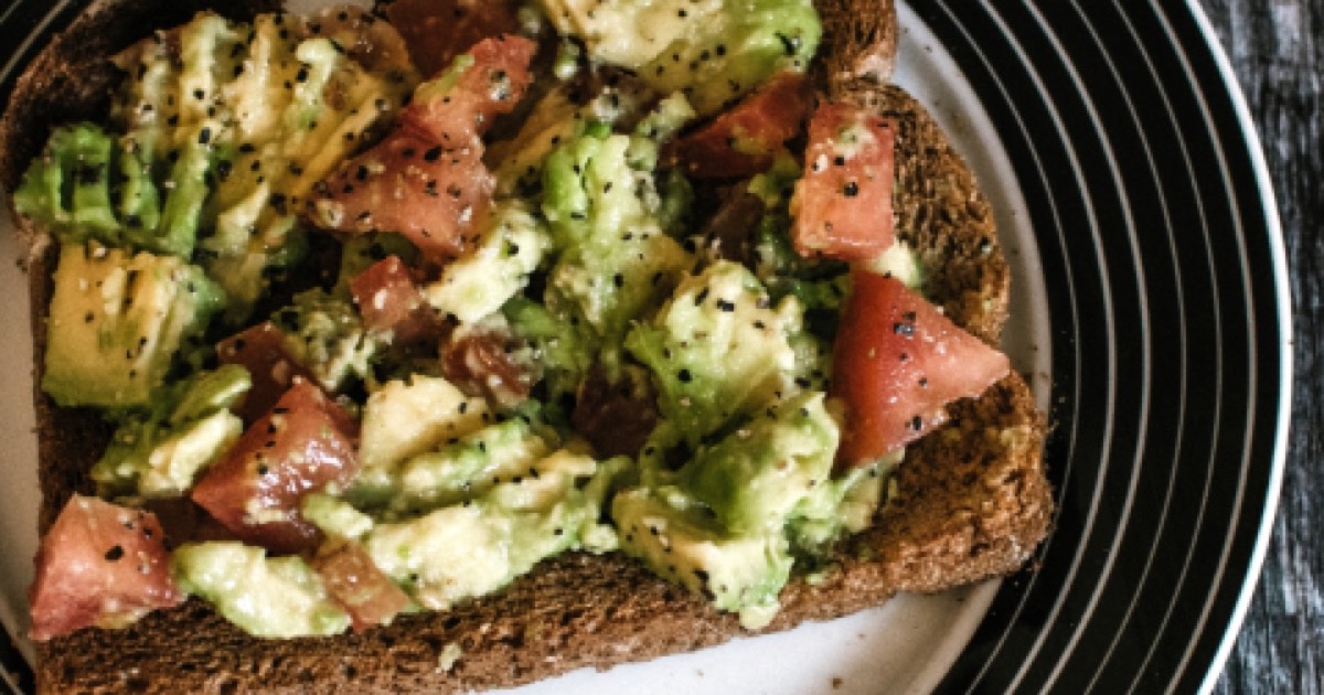 A slice of wholewheat toast topped with mashed avocado, chopped tomato and black seeds, on a plate with a striped rim.